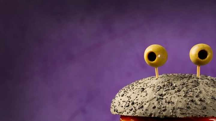 Hamburguesas con pan color negro, vegetales, carne y queso, con aceitunas en la superficie haciendo alusión a monstruos