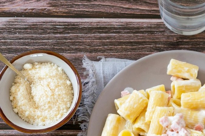 Plato gris con pasta rigatoni con crema y panceta, servido en una mesa junto a un vaso de agua y cubiertos