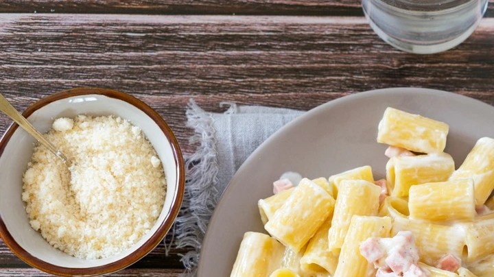 Plato gris con pasta rigatoni con crema y panceta, servido en una mesa junto a un vaso de agua y cubiertos