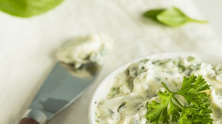 Recipiente blanco con mantequilla de ajo, decorada con hierbas sobre una mesa blanca