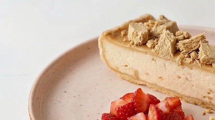 Halva cheesecake sobre un plato rosado, fresas picadas y una cuchara con un trozo de tarta